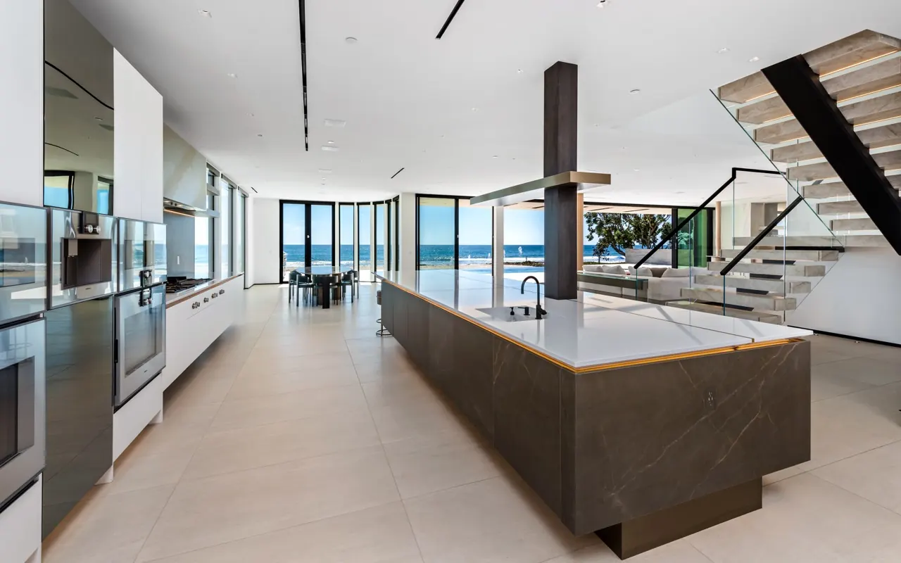 The kitchen: a long stone island under a floating stair, with the dining room and ocean beyond