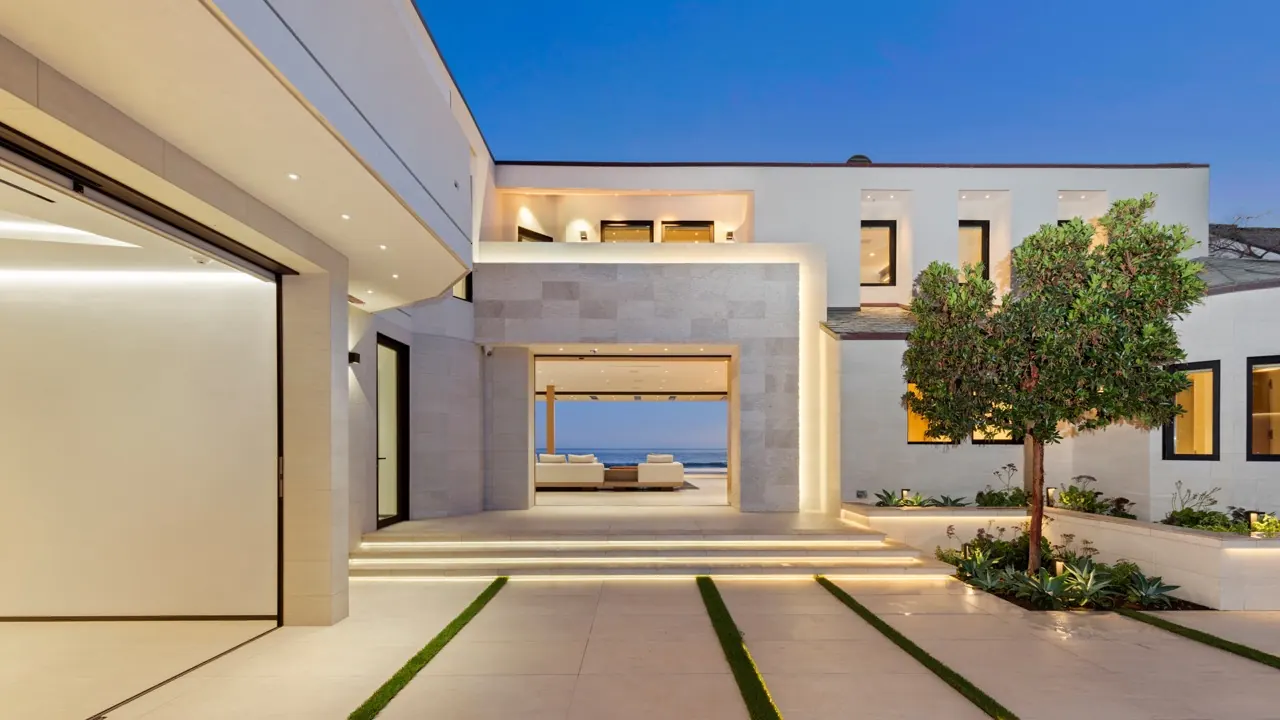 The entry court at dusk: an olive tree, lit steps and a framed view straight through the house to the ocean