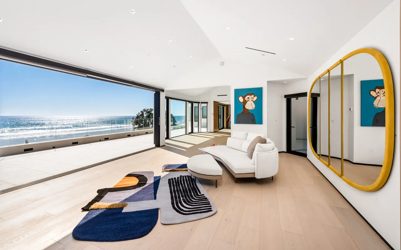 An upstairs sitting room with a sculptural rug and a gold-framed mirror, its glass wall open to the ocean