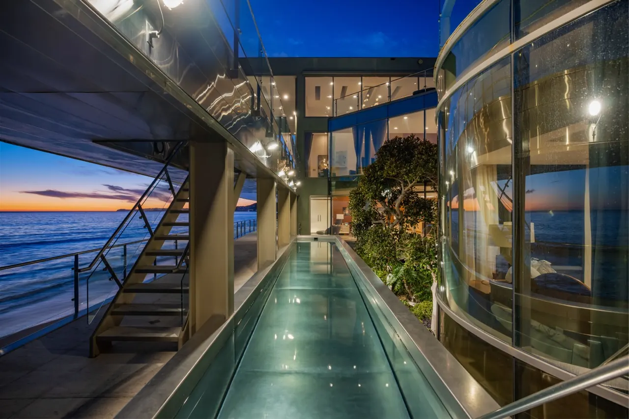The side walkway at dusk, a lit glass-floored pool running between the curved glass stair tower and the ocean