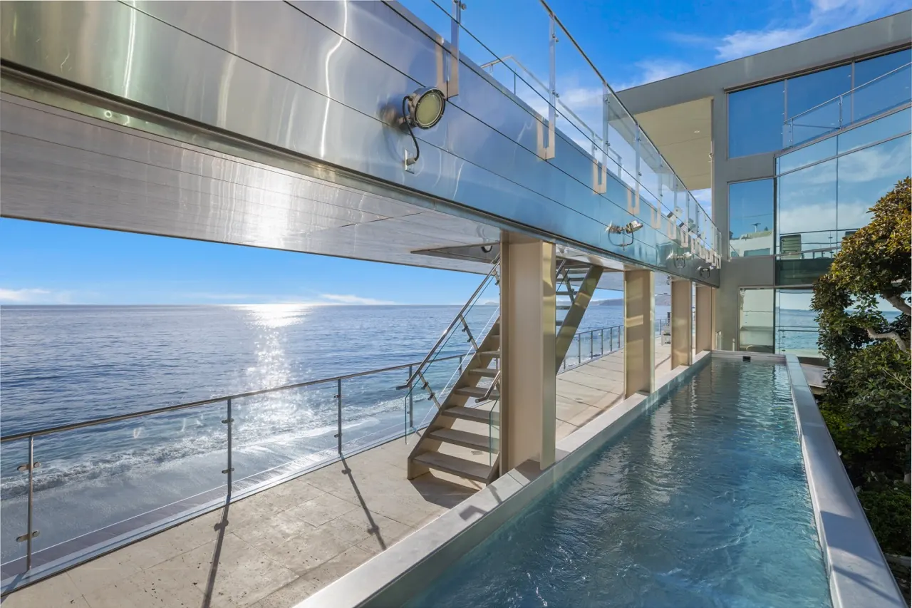 The upper walkway beside a glass-sided lap pool, with stairs dropping to the beach terrace
