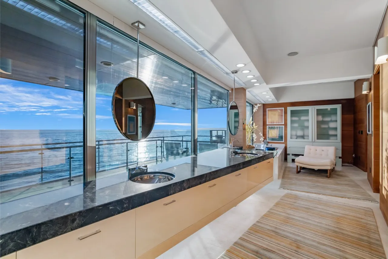 The primary bath: a long stone vanity under a round mirror hung against the ocean glazing