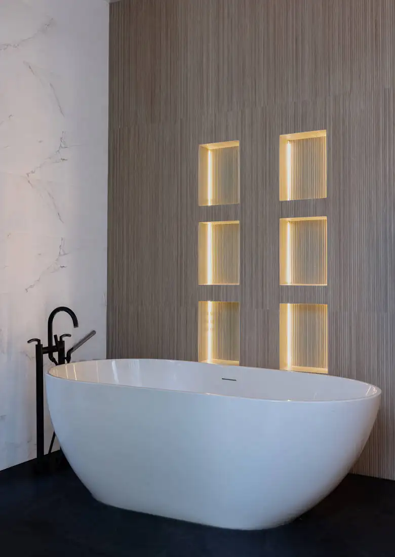 A primary bathroom with a freestanding tub against a marble wall with backlit niches