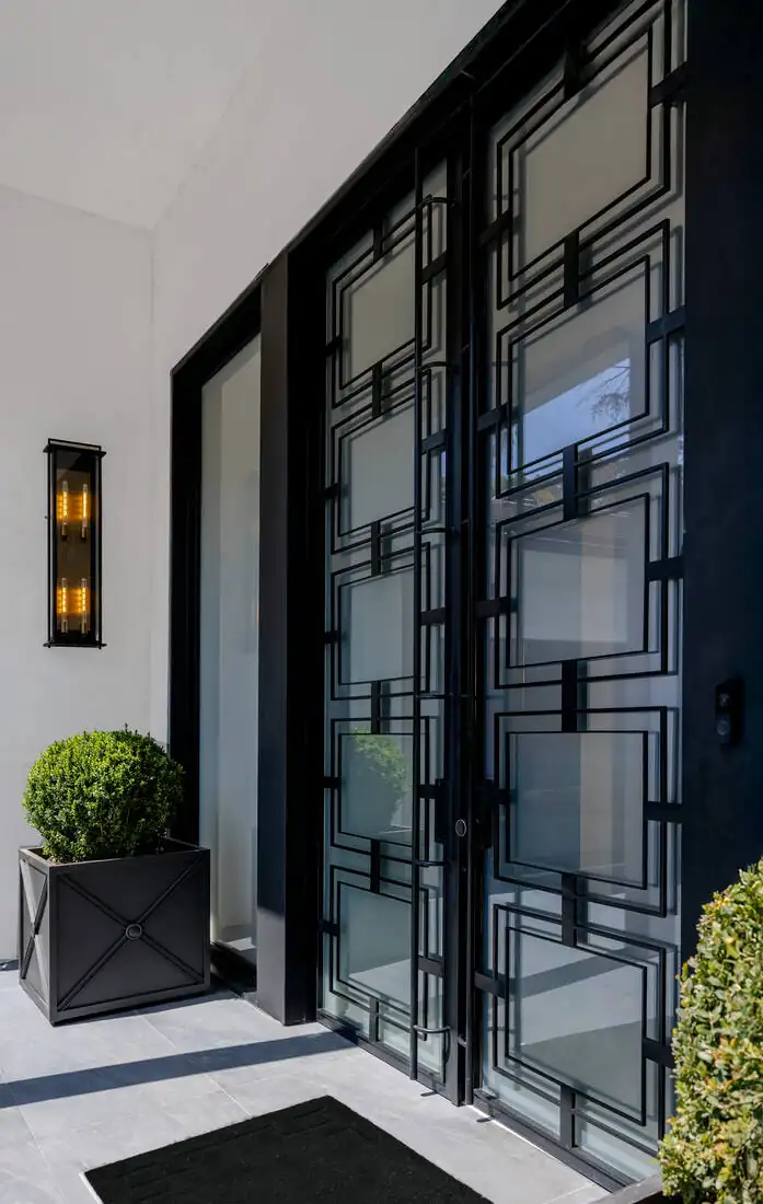 Full-height black steel-and-glass entry doors seen from outside, with a wall lantern and clipped topiary