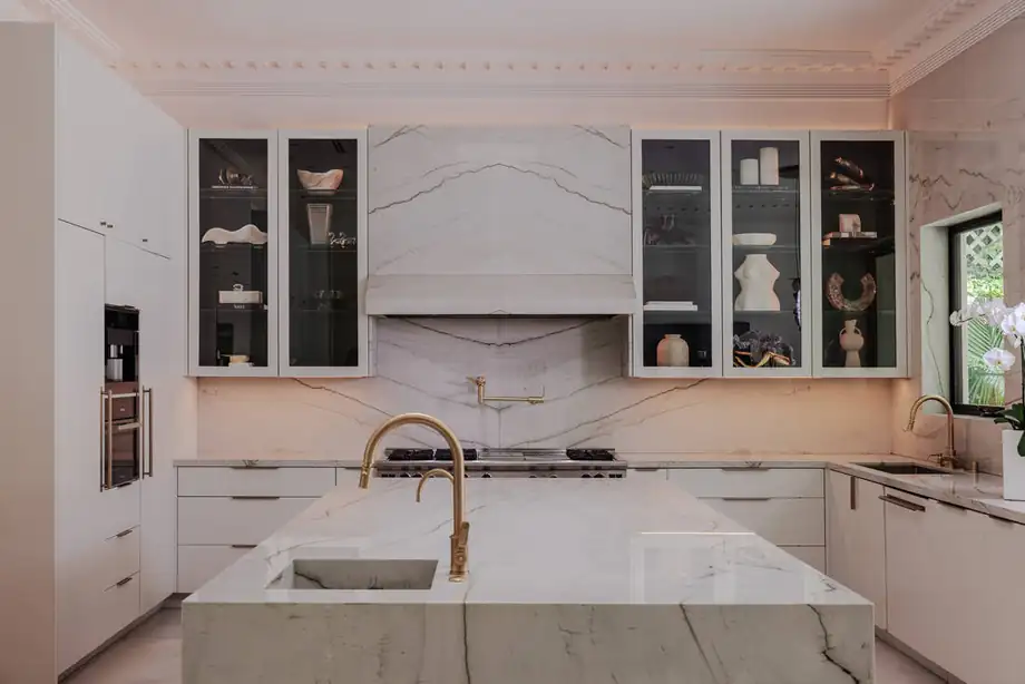 The kitchen after work: white cabinetry, a book-matched marble hood and slab island with a brass tap