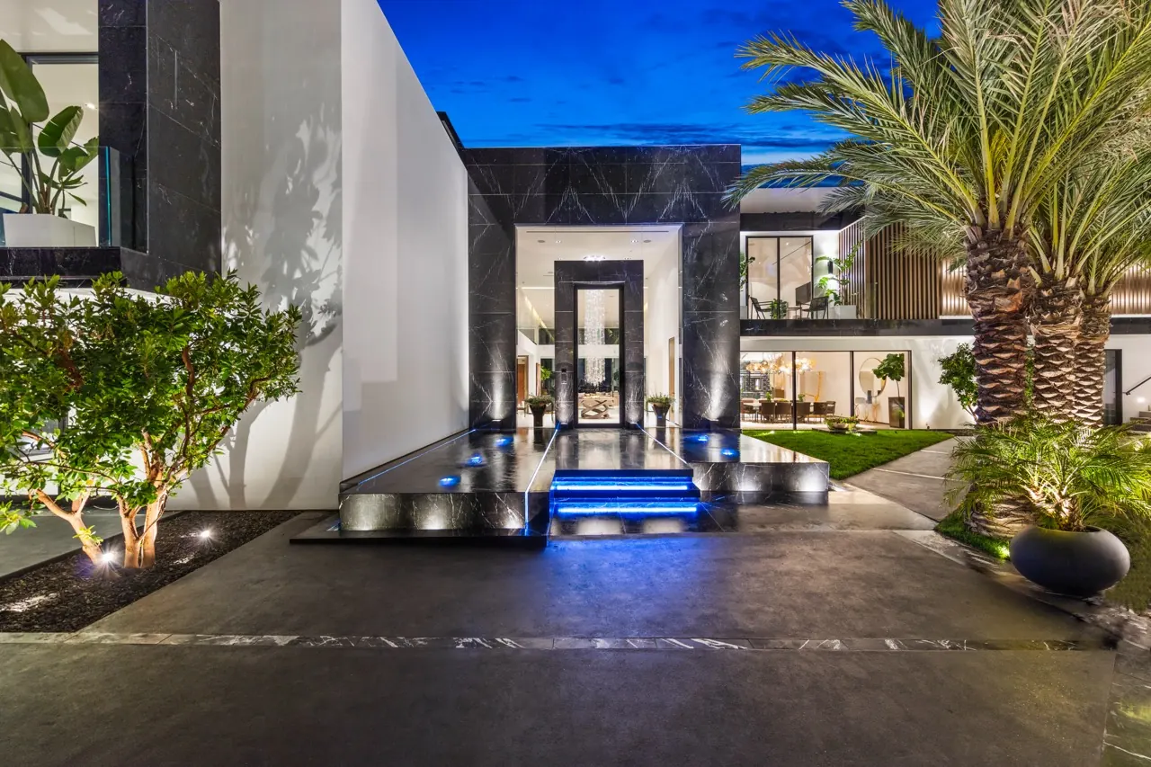 The entry court at dusk: a lit water step leads between dark stone walls and palms to the front door