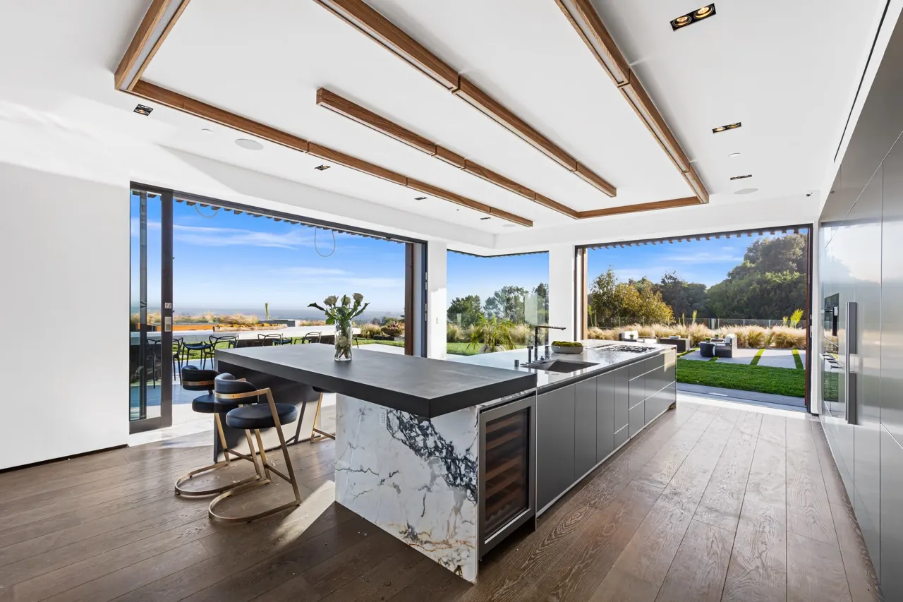 The kitchen: a veined marble waterfall island under timber ceiling beams, with glass walls open to the lawn